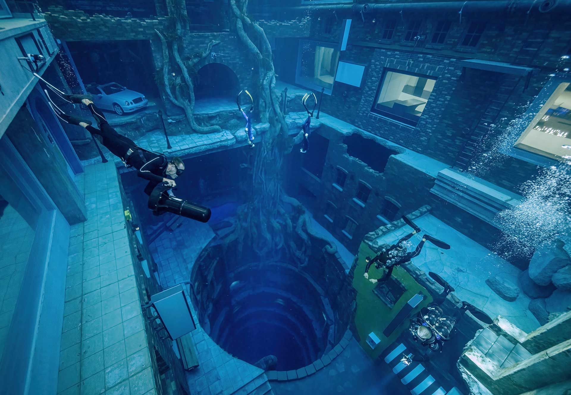 Underwater complex with divers exploring submerged buildings and a deep abyss surrounded by large tree roots.